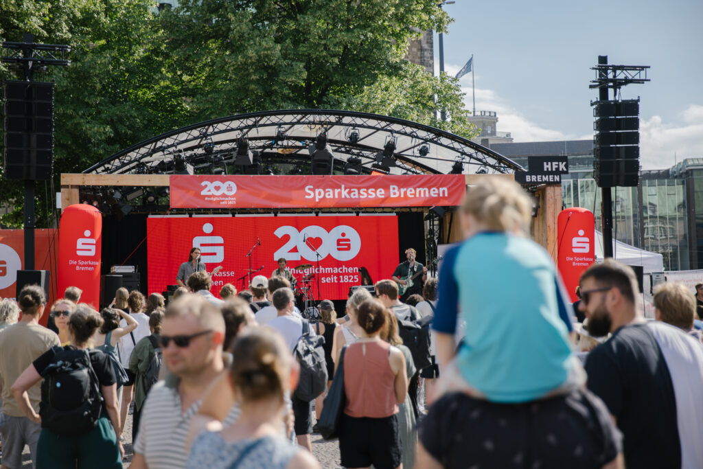 Drei Männer spielen an ihren Instrumenten auf einer Open Air Bühne, die mit roten Sparkassen-Schriftzügen geschmückt ist. Vor der Bühne ist unscharf das Publikum zu erkennen.