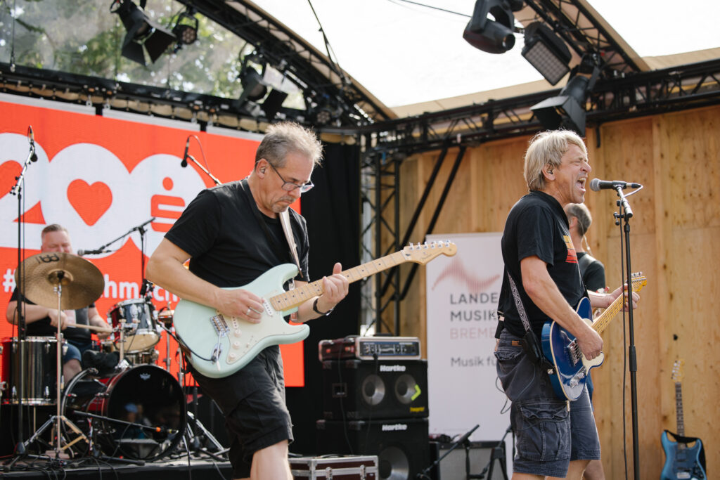 Zwei Männer spielen an ihren Instrumenten auf einer Open Air Bühne, einer der Männer singt leidenschaftlich ins Mikrofon. Im Hintergrund sind zwei weitere Männer unscharf zu erkennen.
