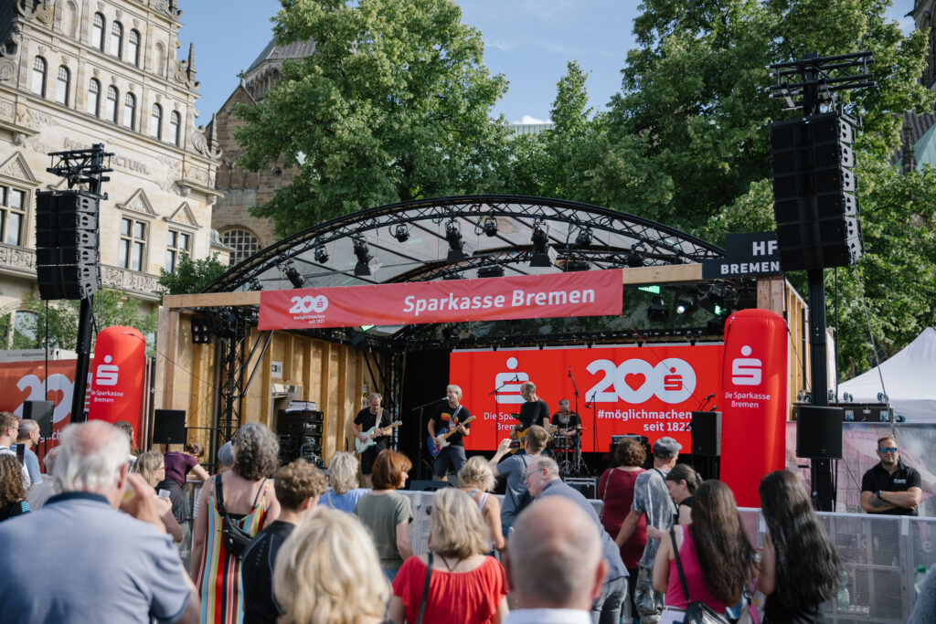 Vier Männer spielen an ihren Instrumenten auf einer Open Air Bühne, die mit roten Sparkassen-Schriftzügen geschmückt ist. Vor der Bühne steht Publikum.
