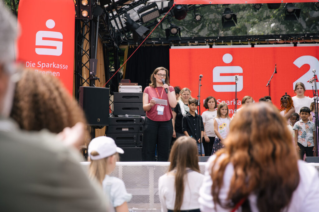 Eine Gruppe bunt gekleideter Kinder steht fröhlich auf einer Open Air Bühne. Seitlich steht eine Frau mit mittellangen braunen Haaren und spricht in ein Mikrofon. Unscharf ist im Vordergrund das Publikum zu erkennen. Die Bühne ist mit roten Sparkassen-Schriftzügen geschmückt.