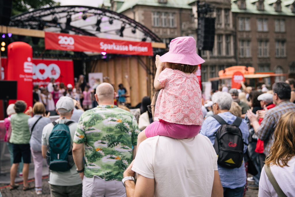 Man sieht viele Menschen von hinten vor einer Open-Air Bühne stehen. Ein rosa gekleidetes Kleinkind sitzt auf den Schultern einer erwachsenen Person und klatscht in die Hände. Die Bühne ist mit roten Bannern der Sparkasse Bremen geschmückt.