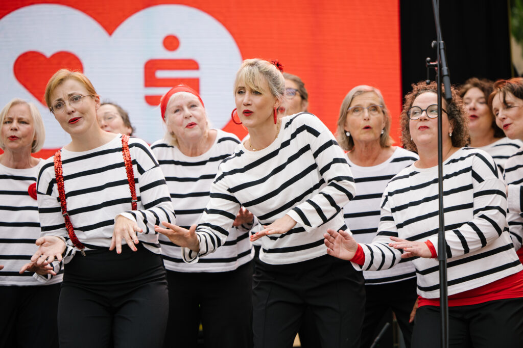 Eine Gruppe Frauen in weiß-schwarz gestreiften Oberteilen, schwarzen Hosen und roten Accessoires stehen singend nebeneinander und bewegen sich zur Musik. Im Hintergrund ist ein rotes Herz und ein rotes Sparkassenzeichen zu sehen.