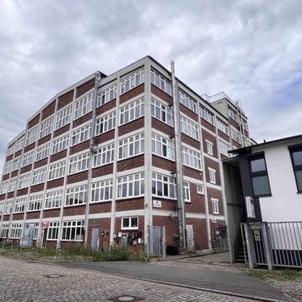 Ein freistehendes großes Gebäude mit fünf Stockwerken aus rotem Backstein und mit weißen Fenstern. Rechts daneben ist ein flacher Bau zu sehen.