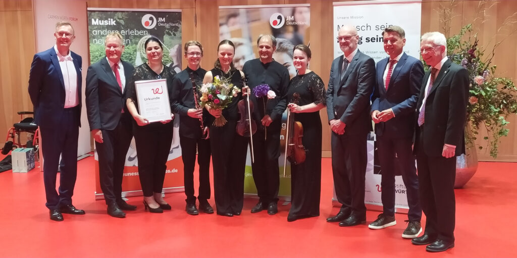 Eine Gruppe von 10 Personen steht nebeneinander auf einem rotem Teppich und vor mehreren Bannern. Sie sind festlich gekleidet und schauen glücklich in die Kamera.