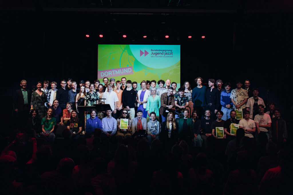 Große Gruppe von zumeist Menschen auf Bühne vor Bildschirm mit Schrift 'Bundesbegegnung Jugend jazzt' und 'Dortmund' in bunter Gestaltung.