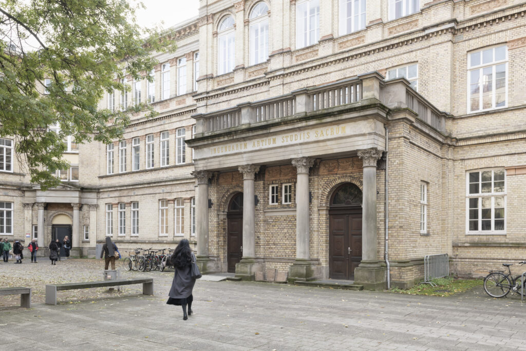 Fassade des historischen Backsteingebäudes der Hochschule für Künste Bremen in der Dechanatstrasse mit Säulenportikus und goldener Inschrift, Personen und Fahrräder davor.