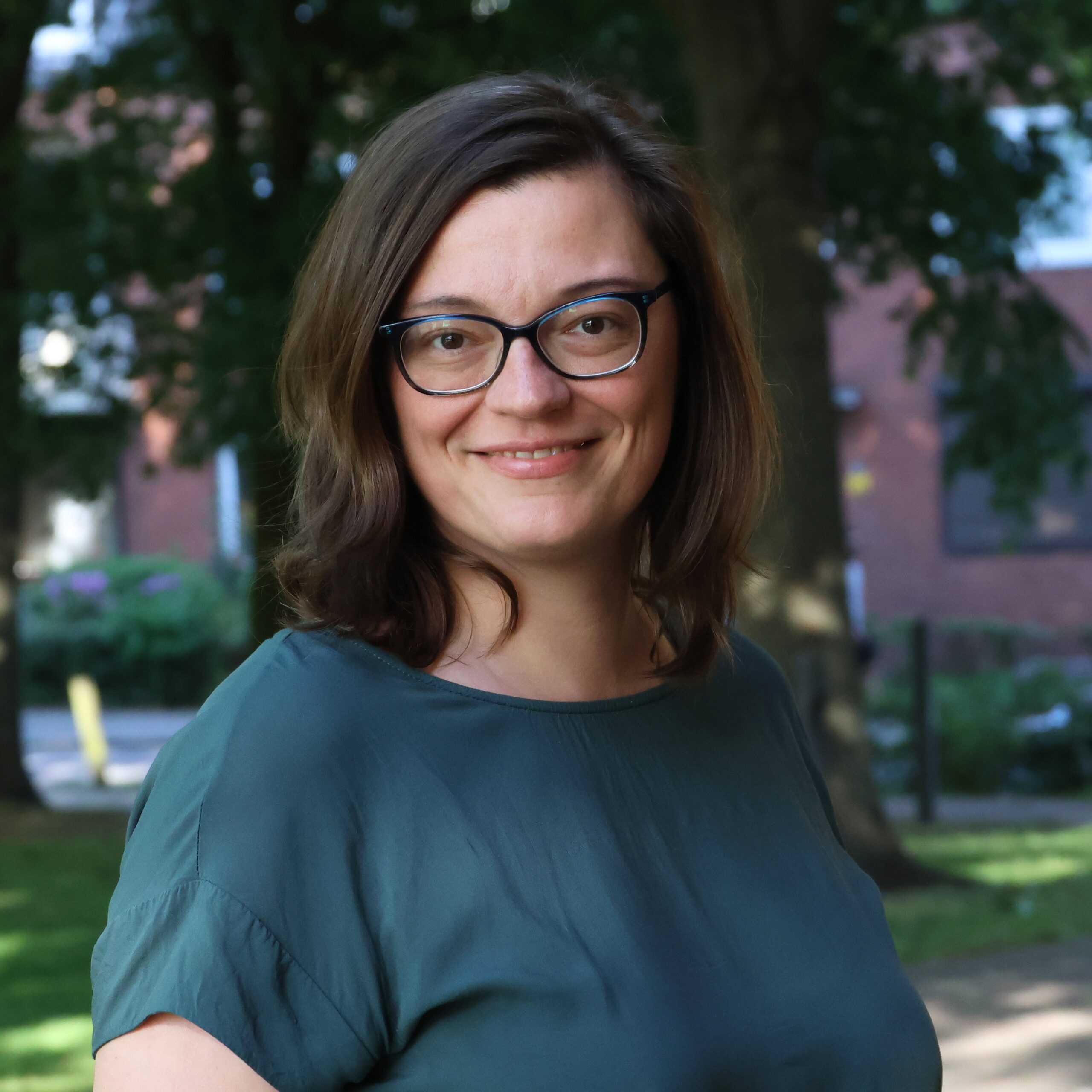 Portrait einer lächelnden brünetten Frau mit mittellangen Haaren, Brille in grüner Umgebung