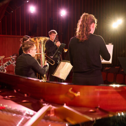 Rücken- und Seitenansicht von vier Jugendlichen mit Blechblasinstrumenten, die durch Scheinwerfer beleuchtet werden. Am linken Rand ist ein Banner mit Aufdruck "Jugend musiziert" zu sehen.