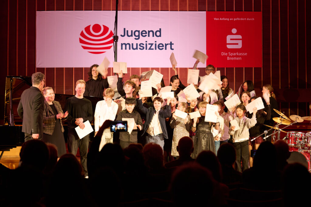 Gruppe von Kindern und Jugendlichen auf einer Bühne hält Urkunden in die Höhe vor einem Banner mit Schriftzug 'Jugend musiziert' und Logo der Sparkasse Bremen.