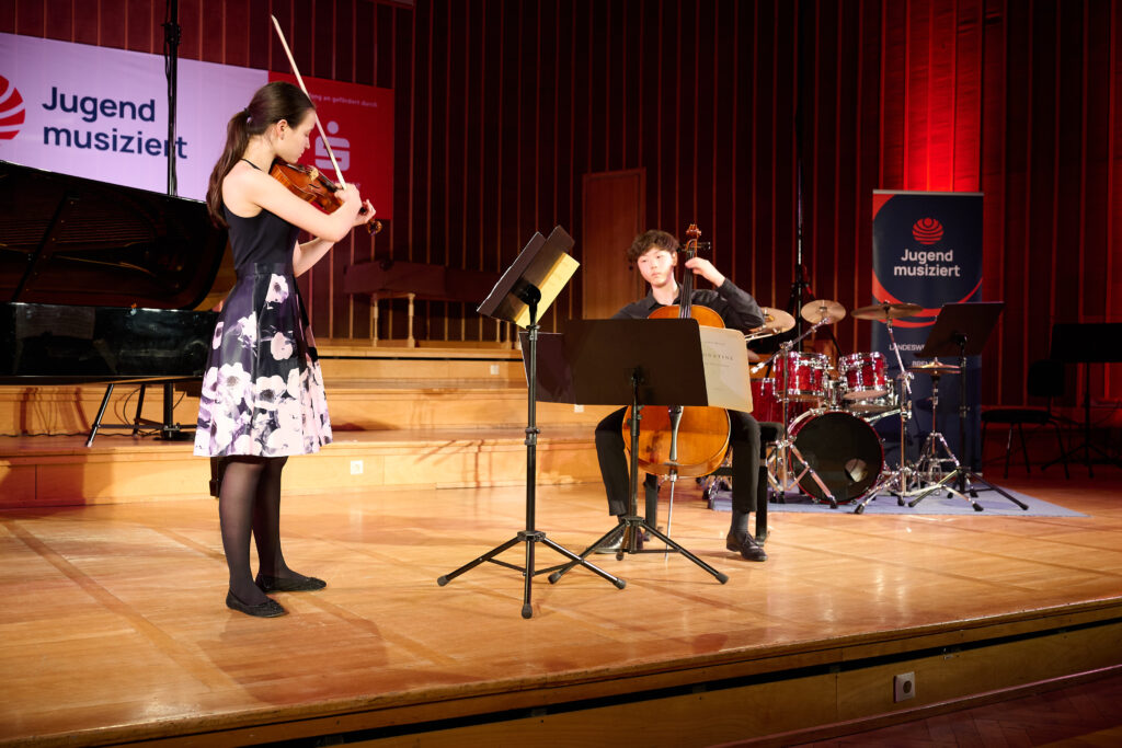 Eine stehende Jugendliche mit Geige und ein sitzender Junge mit Cello musizieren konzentriert auf einer Bühne, vor ihnen stehen Notenständer, im Hintergrund sind ein Drumset und zwei Banner mit der Aufschrift "Jugend musiziert" zu sehen.