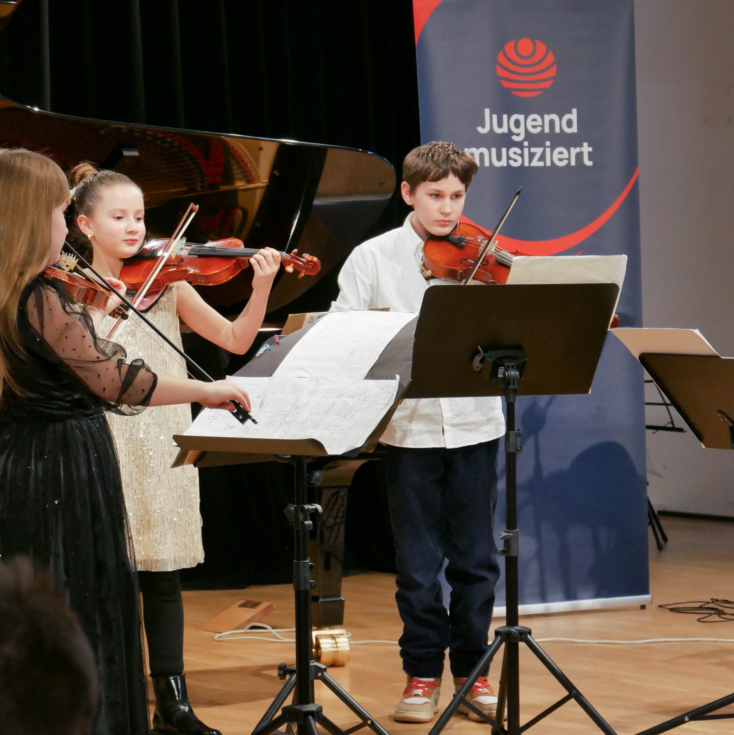 Zwei Mädchen und ein Junge spielen Violinen und Viola. Sie blicken konzentriert in ihre Noten. Im Hintergrund ist ein blaues Banner mit der Aufschrift "Jugend musiziert" zu sehen.