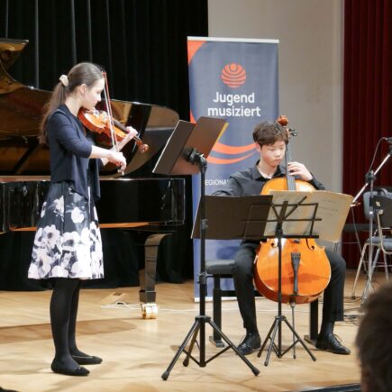 Ein Mädchen und ein Junge spielen Violine und Violoncello, sie blicken dabei konzentriert in ihre Noten. Im Hintergrund ist ein blaues Banner mit der Aufschrift "Jugend musiziert" und ein Teil eines Flügels zu sehen.