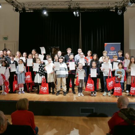 Eine große Gruppe aus Kindern und Jugendlichen steht in mehreren Reihen auf einer Bühne. Sie zeigen ihre Urkunden und halten rot-weiße Sparkassen-Stoffbeutel in den Händen. Sie schauen fröhlich in die Kamera.