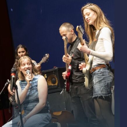 Vier junge Menschen stehen und sitzen auf einer Bühne. Zwei von ihnen spielen E-Gitarren, eine Person hält ein Mikrofon. Die Bühne ist dunkelblau ausgeleuchtet. Die Kleidung der Personen ist lässig und modern.