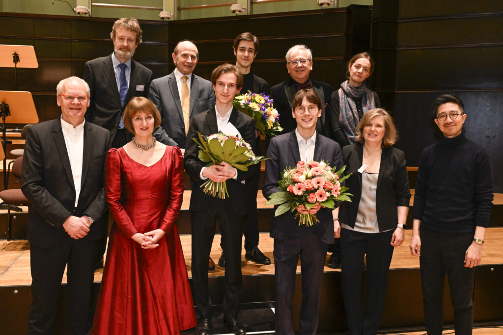 Gruppe von elf Personen in festlicher Kleidung in einem Konzertsaal, drei Personen halten Blumensträuße