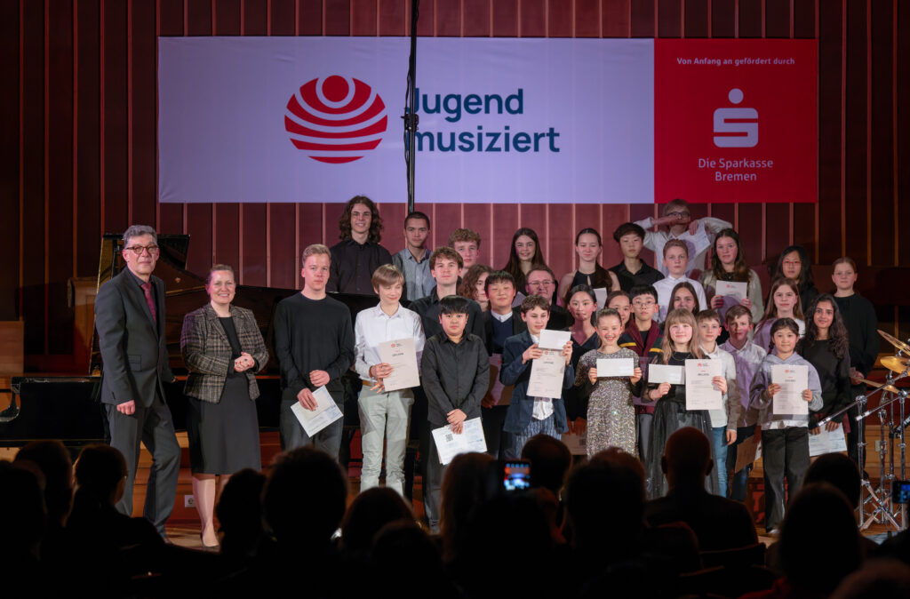 Gruppe von Kindern und Jugendlichen auf einer Bühne hält Urkunden in den Händen vor einem Banner mit Schriftzug 'Jugend musiziert' und Logo der Sparkasse Bremen.