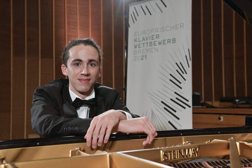 Junger Mann im Anzug lehnt an einem Flügel mit der Aufschrift Steinway vor einem Banner mit der Aufschrift 'EUROPÄISCHER KLAVIER WETTBEWERB BREMEN 2021'