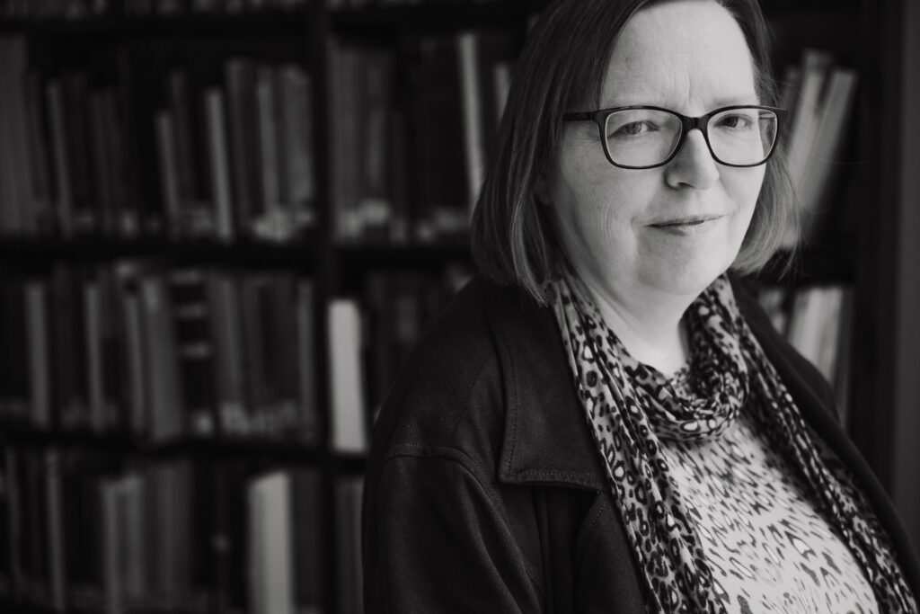 Schwarzweiß-Porträt einer Frau mit Brille und gemustertem Schal vor einem Bücherregal